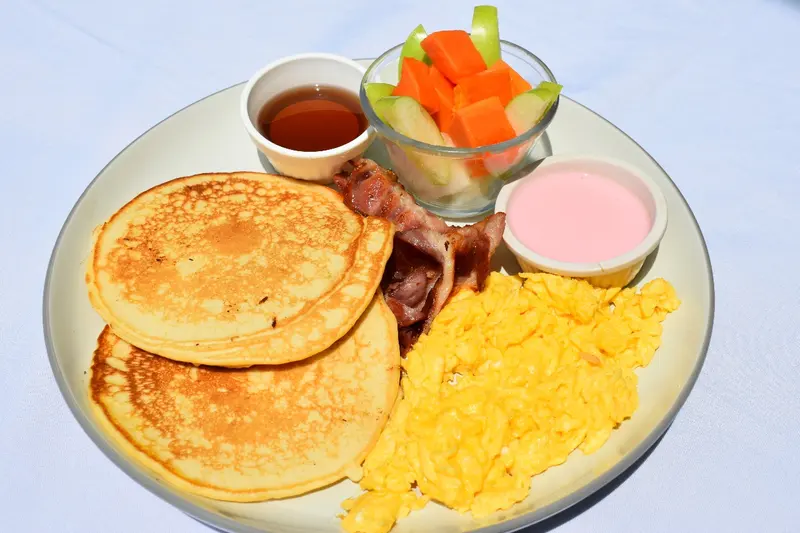 Desayuno con pancakes, huevos revueltos, tocineta, fruta y miel